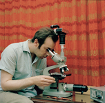 831001 Afbeelding van Peter van der Linden achter een tri-oculair microscoop met bovenop een kleinbeeld fotocamera.